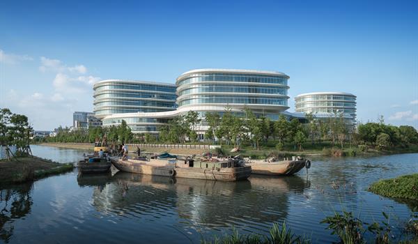 glazed office building with three visible volumes, with rounded oval shapes and curvilinear podium connection, all viewed from across a waterway with boats in the foreground